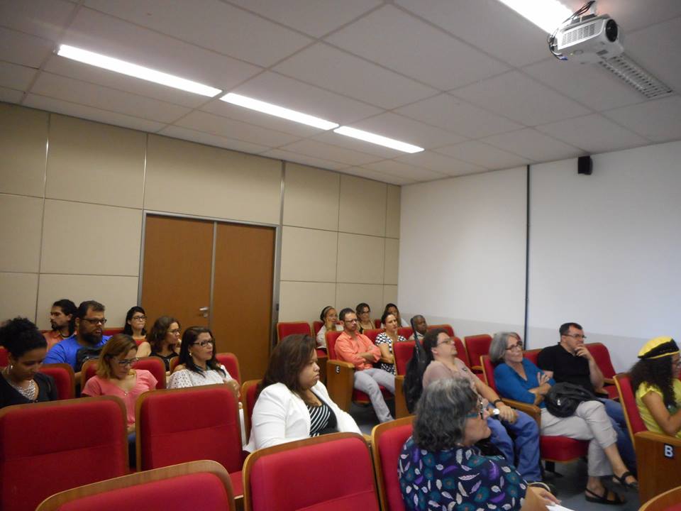 No auditório da Biblioteca Central todas as poltronas são vermelhas, 19 pessoas estão na foto, na primeira fileira, do lado direito tem uma mulher sentada, ela está usando uma blusa amarela e boina amarela com uma faixa preta; na segunda fileira tem um homem de blusa preta e calça jeans e a sua esquerda uma mulher com blusa azul e calça bege, a esquerda, no corredor, um homem cadeirante vestindo uma blusa marrom e calça jeans; a terceira fileira está vazia; na quarta fileira tem um homem com uma blusa bege, a sua esquerda uma mulher usando um vestido quadriculado preto e branco, a sua esquerda um homem usando blusa salmão e calça bege; na quinta, e última fileira, vestindo branco, a sua esquerda uma mulher vestindo preto e a sua esquerda uma mulher vestindo bege. Do lado esquerdo do auditório, na primeira fileira uma mulher está usando uma blusa azul escura com estampa de penas roxas, brancas e verdes; na segunda fileira uma mulher utilizando uma blusa preta com listras brancas e um casaco branco; na terceira fileira uma mulher com vestido branco, a sua esquerda uma mulher com blusa rosa e calça jeans e a sua esquerda uma mulher com blusa preta, na quarta fileira uma mulher com blusa preta e a sua esquerda um homem com blusa azul; e na quinta, e última fileira, uma mulher com blusa vermelha e a sua esquerda um homem com blusa salmão.